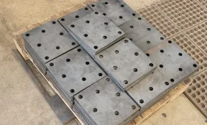 Una pila de placas de metal con agujeros espaciados uniformemente, colocadas sobre una plataforma de madera, situada junto a una alfombra de goma con patrones circulares.