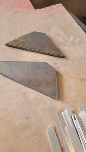 Dos piezas triangulares de metal con una superficie oxidada yacen sobre un banco de trabajo al lado de tiras de metal cuidadosamente apiladas.