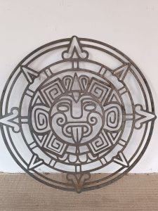 Arte de pared de metal que presenta un diseño circular de inspiración azteca con patrones geométricos y una cara estilizada en el centro.