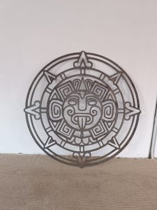 Una pieza de arte circular de metal con patrones geométricos y un diseño de cara central cuelga en una pared blanca.