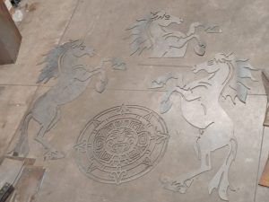 Recortes de metal de tres caballos encabritados y un diseño circular sobre un piso de concreto.