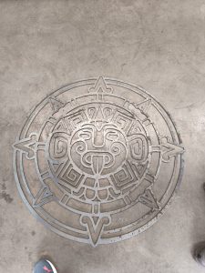 Diseño metálico de una cara de sol estilizada en un patrón circular, que recuerda a antiguos motivos aztecas o mayas, sobre un suelo de hormigón.