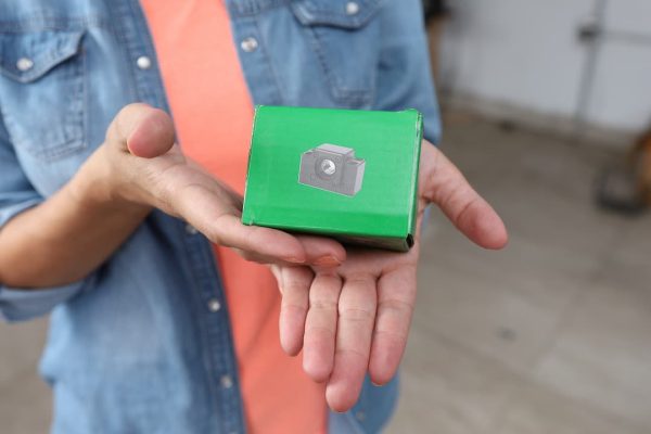 Persona con una camisa vaquera sosteniendo una pequeña caja verde con un icono de cámara encima.