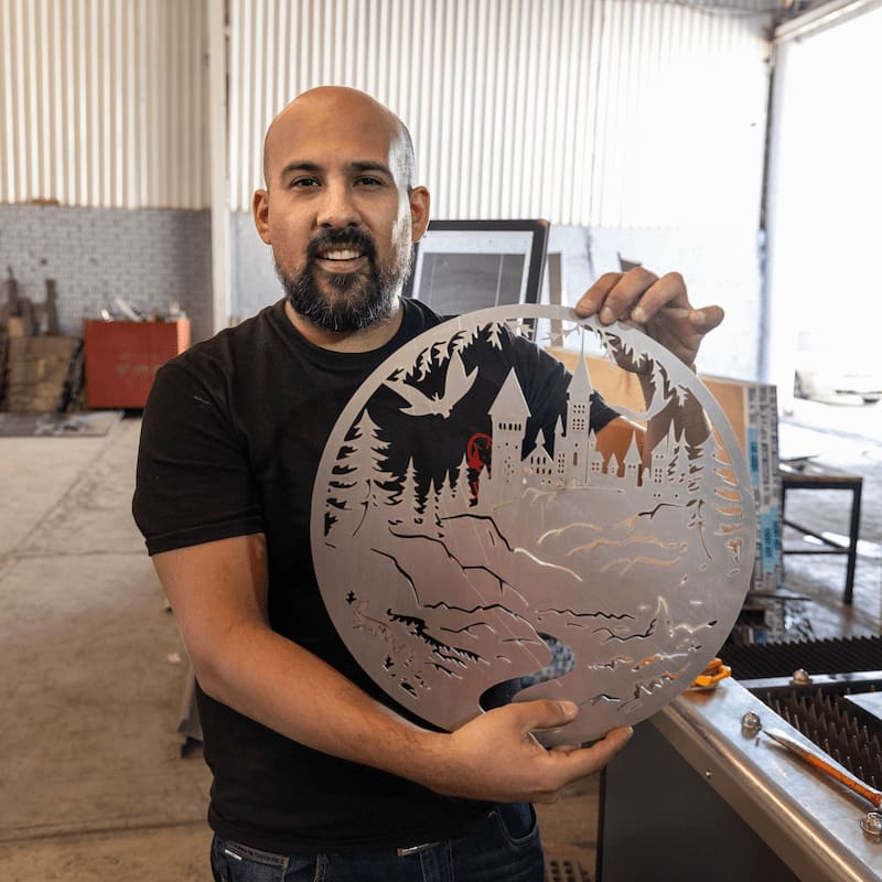 Un hombre en un taller sostiene una pieza de arte redonda de acero inoxidable cortada con un laser cnc fibra que representa una escena de montaña y bosque con un castillo.