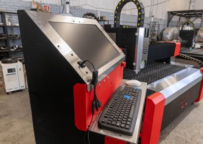 Máquina industrial con pantalla táctil, teclado y cables, situada en un taller con estanterías y diversos equipos al fondo.