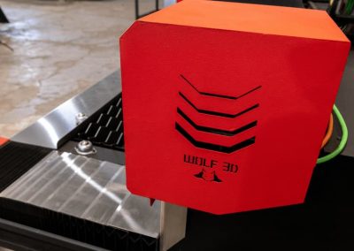 Primer plano de un componente rojo de impresora 3D de Wolf 3D, con recortes geométricos y un logotipo con dos lobos. La precisión del láser CNC de fibra añade detalles, sobre un suelo de hormigón texturizado.