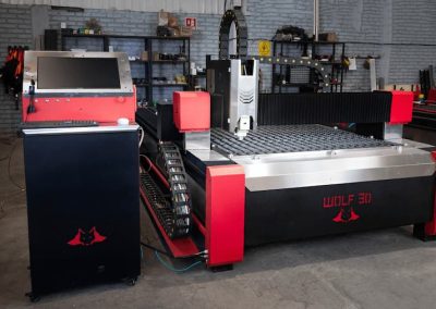 Una máquina laser en un espacio de trabajo industrial con un panel de control a la izquierda. Estantes con herramientas y materiales alinean la pared de ladrillos del fondo.