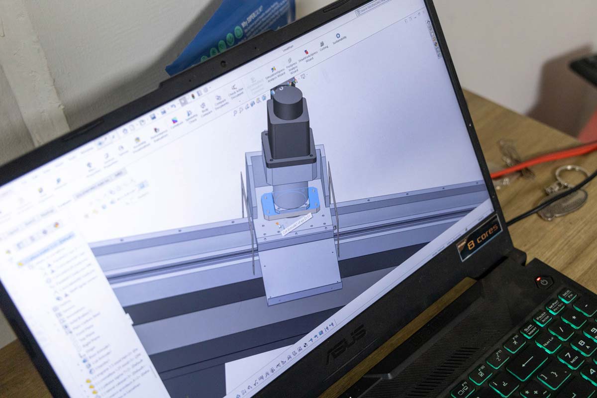 Una computadora portátil muestra un modelo CAD 3D de un componente mecánico en su pantalla, con un software de diseño láser CNC abierto y varias barras de herramientas visibles.