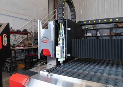 Una vista de primer plano de una máquina láser CNC industrial en un taller, que muestra su cabezal de corte y su superficie de trabajo de metal, con herramientas y estantes en el fondo.