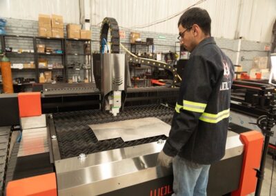Un hombre maneja una máquina de corte por láser CNC en un taller industrial, con estanterías de equipos y suministros al fondo.
