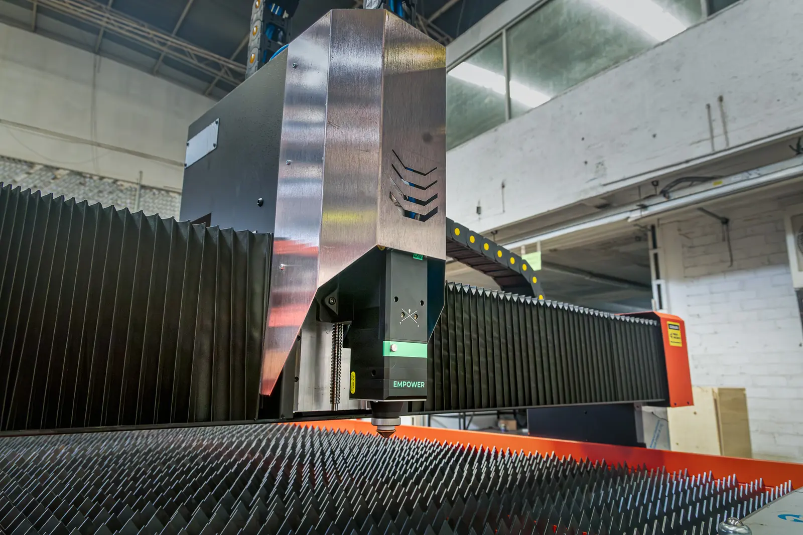 Primer plano de una máquina industrial de corte por láser en funcionamiento, situada en un entorno fabril con rejillas metálicas y una iluminación cenital intensa.