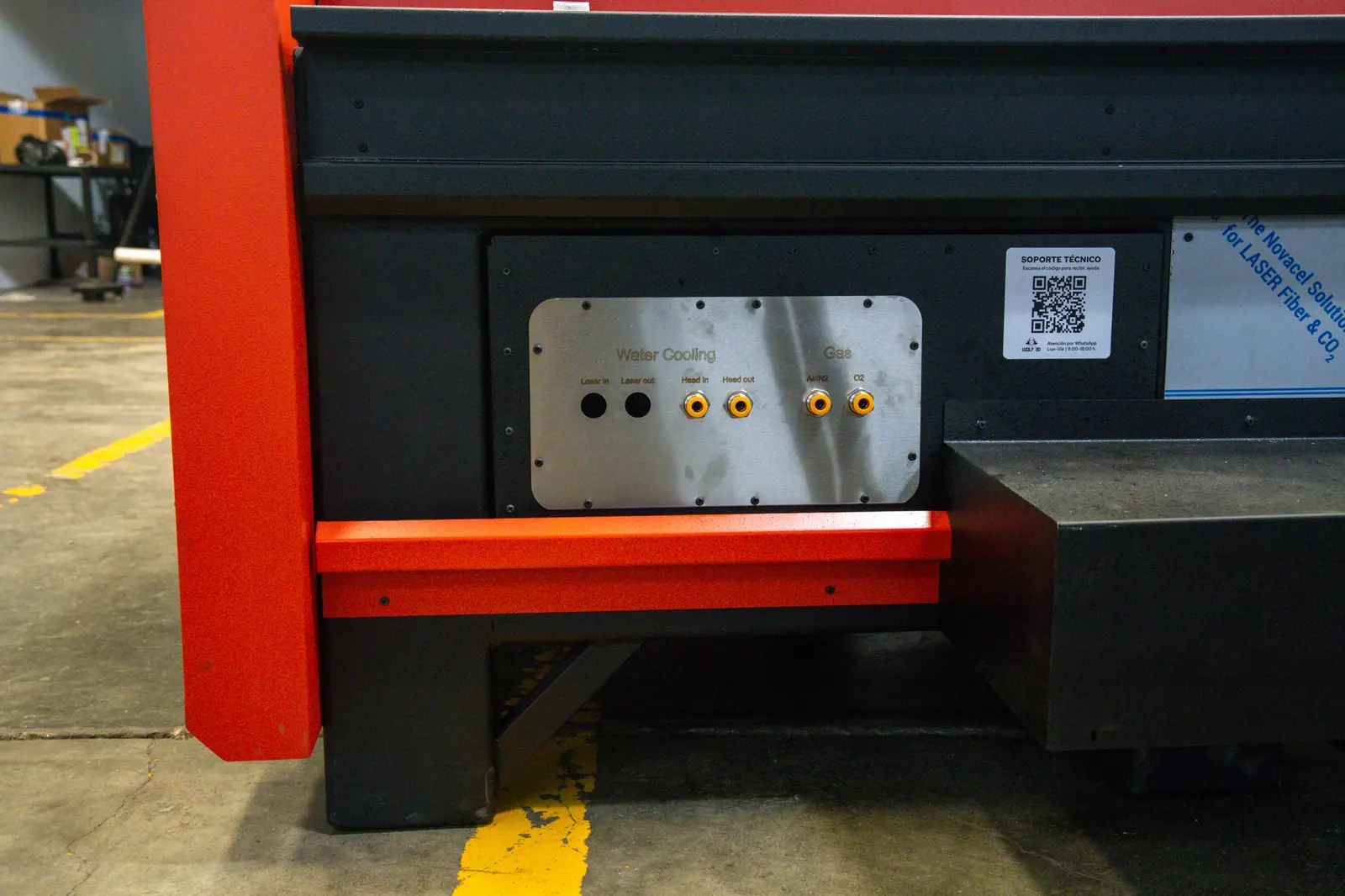 Primer plano de un panel de una máquina en el que se distinguen las tomas de refrigeración por agua y de gas, un código QR, y, al fondo, el suelo industrial y el equipamiento.