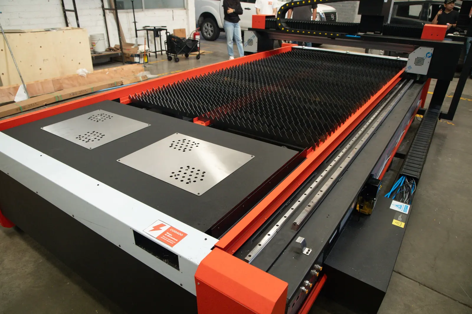 Gran máquina industrial de corte por láser CNC con un bastidor rojo y negro en un entorno de taller, que cuenta con una plataforma con púas para la colocación del material y cubiertas metálicas con orificios de ventilación.