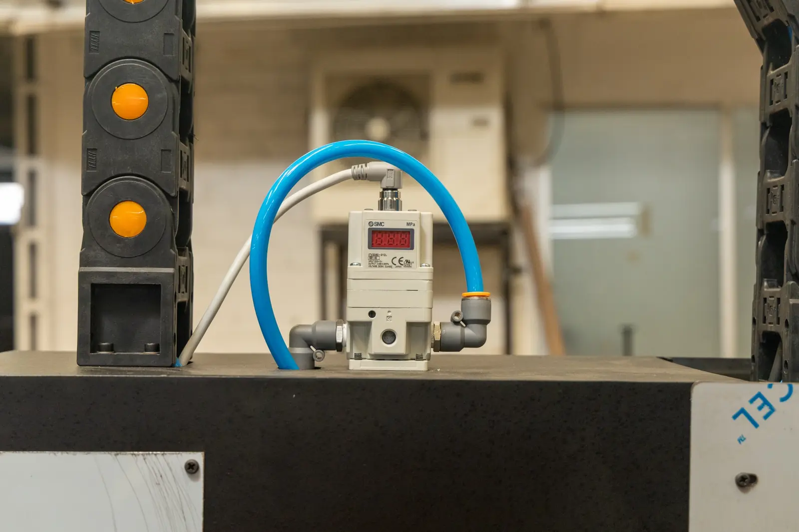 Un regulador de presión digital con una manguera azul está instalado sobre una superficie negra en un entorno industrial. A la izquierda se ven unos botones amarillos.