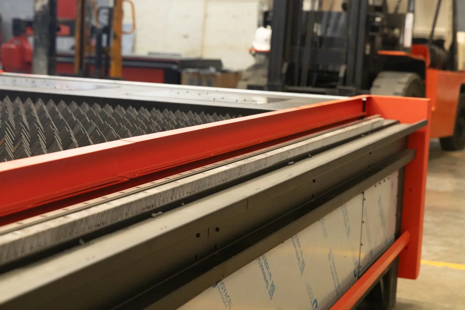 Primer plano de maquinaria industrial en una fábrica, en el que se aprecia un borde metálico rojo y una plataforma con forma de diente de sierra, con una carretilla elevadora y un trabajador difuminados al fondo.