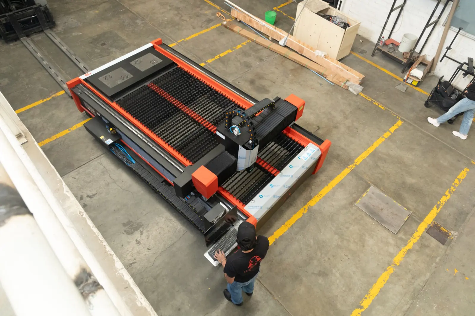 Una persona maneja una gran máquina industrial CNC en una fábrica, donde el suelo de hormigón está marcado con líneas amarillas de seguridad.