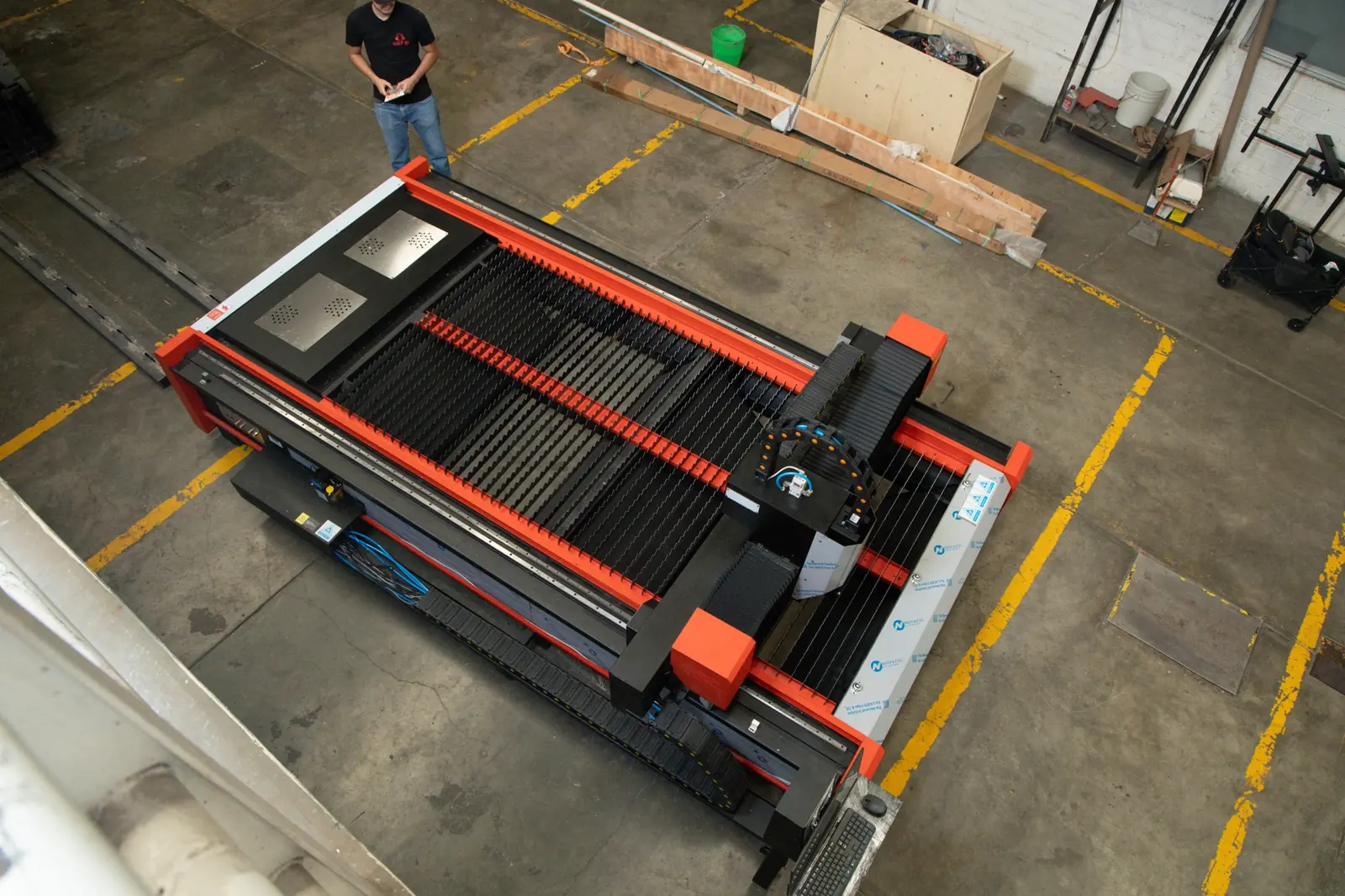 Una persona se encuentra de pie junto a una gran máquina CNC industrial en el interior de una fábrica, vista desde arriba. La máquina es de color rojo y negro, con una base metálica y un panel de control.